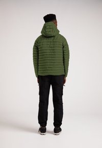 Olivgrön pufferjacka med huva, med horisontell quiltning. Buren med svarta cargobyxor och sportskor, sedd bakifrån.