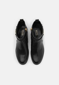 Bottes basses en cuir noir avec une finition lisse et élégante et des accents de ferrures dorées à la cheville, présentant un design minimaliste.