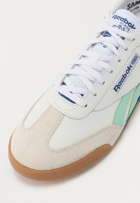 Fehér bőr sportcipő velúr orral, türkiz kiegészítőkkel és kék részletekkel. Fehér fűzőkkel és gumi talppal rendelkezik. A Reebok logó látható rajta.
