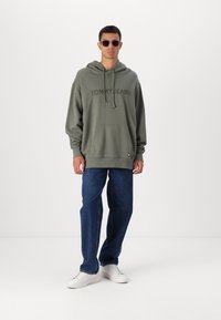 Felpa verde oliva con ricamo "TOMMY JEANS", tasca frontale, indossata con jeans blu e sneakers bianche, completata da occhiali da sole tondi.