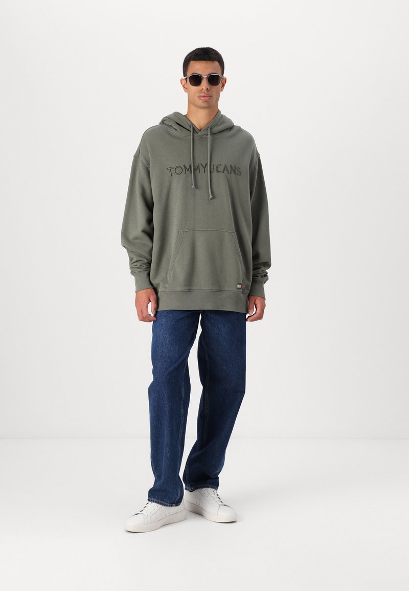 Felpa verde oliva con ricamo "TOMMY JEANS", tasca frontale, indossata con jeans blu e sneakers bianche, completata da occhiali da sole tondi.