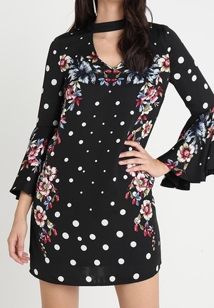 Robe noire à pois blancs, motifs floraux colorés, manches évasées et col choker portée par une personne debout.