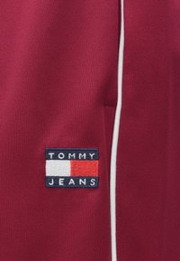 Bordo spalvos sportinės kelnės su baltu apvadu, ant stačiakampio lipduko išsiuvinėtu "TOMMY JEANS" logotipu mėlynos, baltos ir raudonos spalvų.