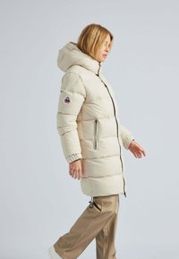 Manteau matelassé beige clair avec capuche, doté d'une fermeture éclair à l'avant, de deux poches latérales et de poignets élastiques. Comprend un patch logo sur la manche.