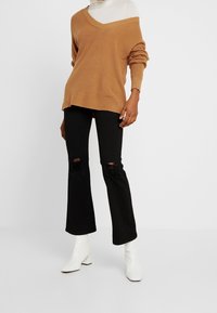 Przesadnie duży brązowy sweter dziany z dekoltem w serek i opuszczonymi ramionami, zestawiony z czarnymi rozszerzanymi dżinsami z rozdarciami na kolanach oraz białymi botkami za kostkę.