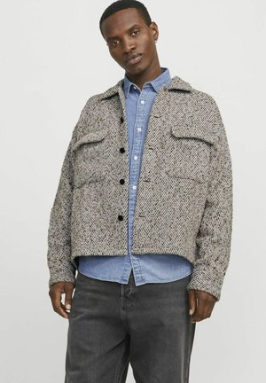 Veste courte texturée grise et marron avec deux poches avant, boutons noirs et col, portée par-dessus une chemise bleu clair.