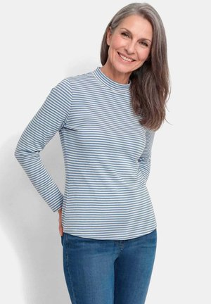 Femme d'âge moyen souriante aux cheveux gris, portant une chemise à manches longues rayée bleu et blanc et un jean bleu, se tient les mains dans le dos.