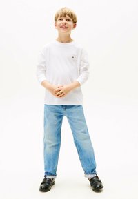 Weißes Langarmshirt mit kleinem Logo, hellblaue Jeans, schwarze Slipper; stehende Pose vor einem hellen Hintergrund.