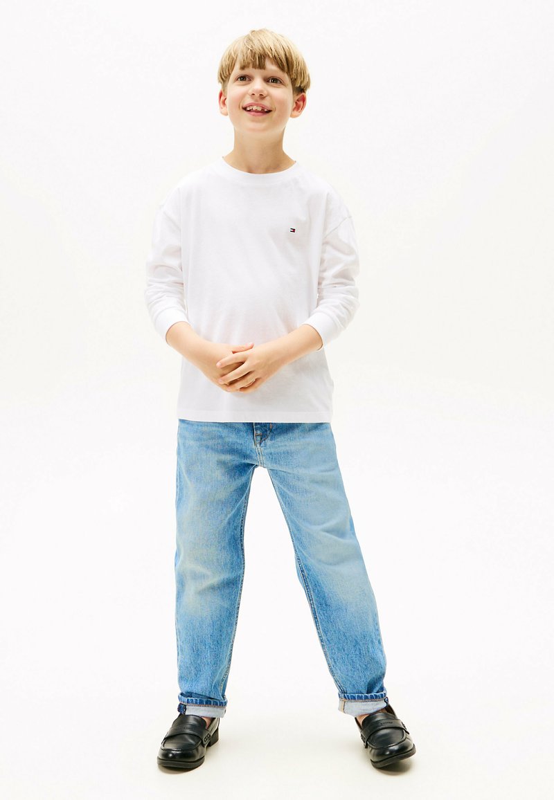 Weißes Langarmshirt mit kleinem Logo, hellblaue Jeans, schwarze Slipper; stehende Pose vor einem hellen Hintergrund.