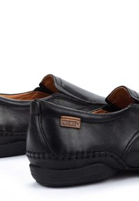 Zapatos de cuero negro sin cordones con una textura suave, que presentan una costura de acento marrón y un pequeño logo de 'Pikolinos' en el talón.