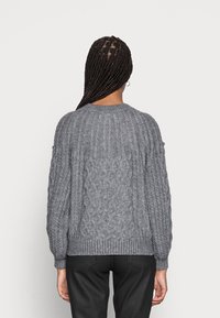 Osoba s dlouhými copy, která nosí šedý svetr se vzorem copu a černé kalhoty, stojící zády k jednoduchému světlému pozadí.