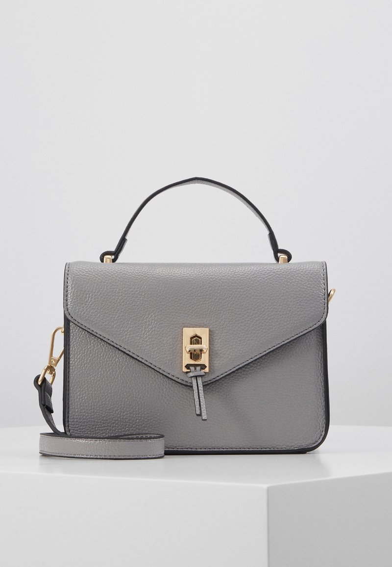Sac à main en cuir gris avec poignée supérieure, fermoir doré et bandoulière amovible, posé sur une surface blanche sur un fond clair.