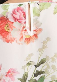 Blouse florale avec de grandes pivoines roses et corail sur un fond doux et clair. Comprend une petite ouverture en forme de serrure à l'arrière avec un bouton.