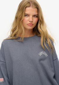 Sweat-shirt gris surdimensionné en coton avec col en côte, arborant un logo "SUPERDRY" et un écusson avec un drapeau américain sur la manche.