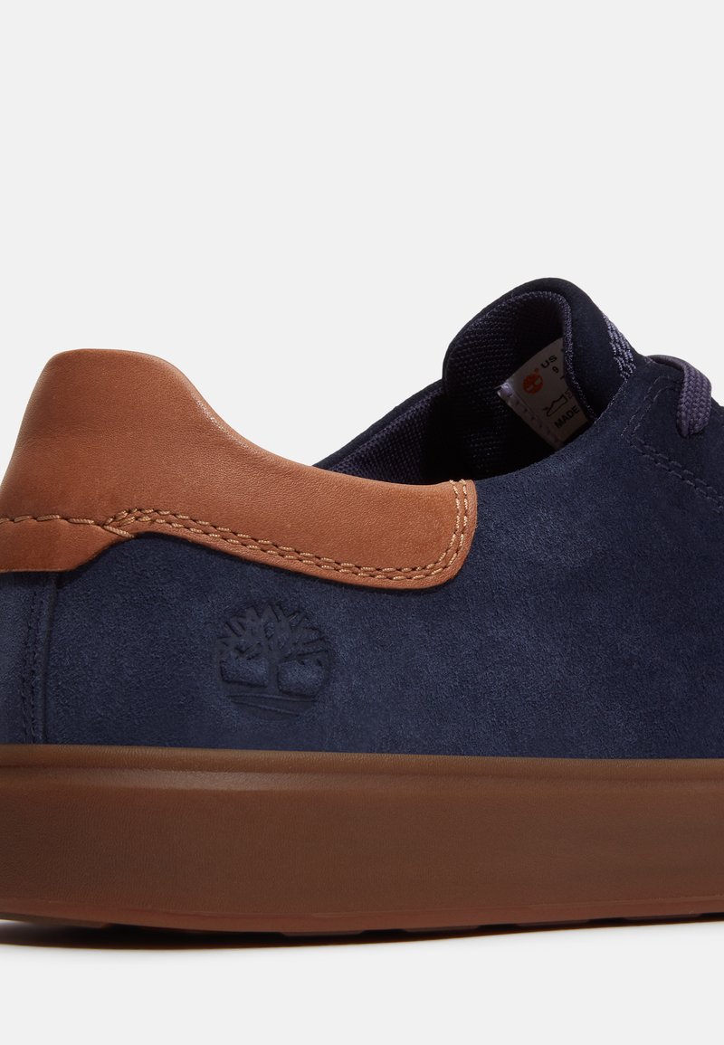 Scarpa scamosciata blu navy con dettagli in pelle marrone sul tallone, suola in gomma e logo impresso. Presenta lacci piatti e colletto imbottito.
