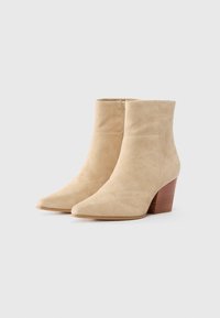 Beige suede ankle boots with a pointed toe, featuring a side zipper, and a stacked, brown wooden heel. Smooth texture and minimalistic design.