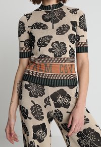 Top de punto con patrones florales negros sobre un fondo beige, cuello con cremallera y dobladillo a rayas con "PALM COVE" en tonos tierra.
