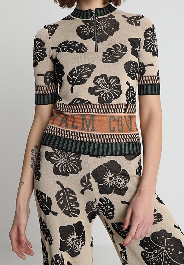 Top de punto con patrones florales negros sobre un fondo beige, cuello con cremallera y dobladillo a rayas con "PALM COVE" en tonos tierra.