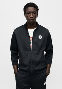 Μαύρο μπουφάν με φερμουάρ και όρθιο γιακά, αθλητικό σχέδιο, με λογότυπο σε ύφασμα στο στήθος, και ταιριαστό παντελόνι με φερμουάρ στα πλάγια.