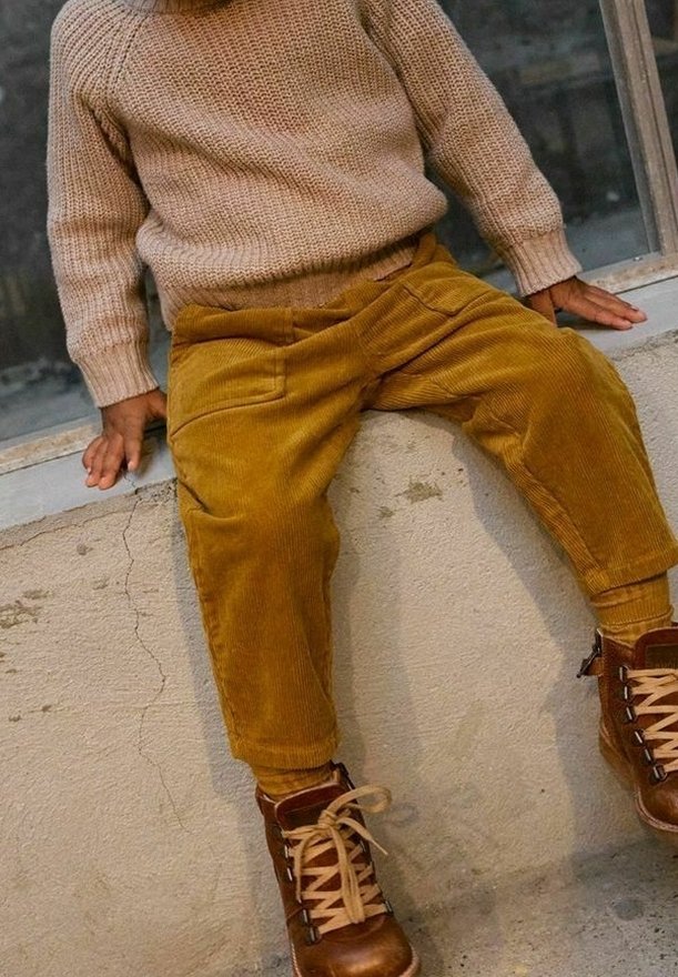 Enfant portant un pantalon en velours côtelé moutarde, un pull en tricot beige et des bottines marron à lacets, assis sur un rebord en béton les mains posées à côté.