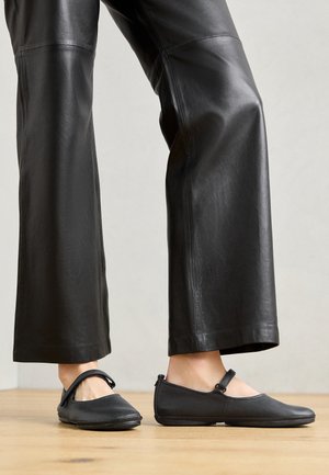 Schwarze Lederflache Schuhe mit runder Zehenform, ausgestattet mit einem Knöchelriemen. Kombiniert mit lockeren schwarzen Lederhosen, vor einem hellen Hintergrund.