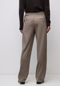 Osoba nosząca taupe'owe spodnie z szerokimi nogawkami, elastycznym paskiem w talii i klasycznymi kieszeniami z tyłu, w połączeniu z ciemnym topem z długim rękawem oraz czarnymi butami.