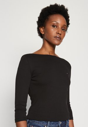 Femeie cu păr scurt și creț, purtând un top negru cu mâneci trei sferturi și blugi albaștri, privind înainte pe un fundal simplu și deschis.