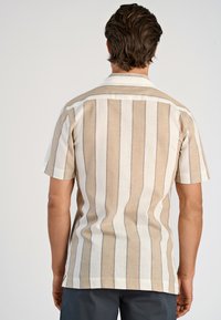 Man in een beige en wit gestreepte short-sleeve shirt met donkere broeken, gezien vanaf de achterkant tegen een effen achtergrond.