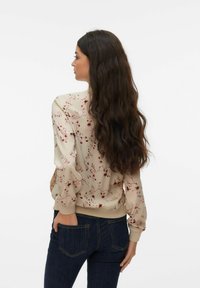 Femme aux longs cheveux noirs portant un chemisier beige à fleurs et un jean bleu foncé, debout de dos avec une main dans la poche.