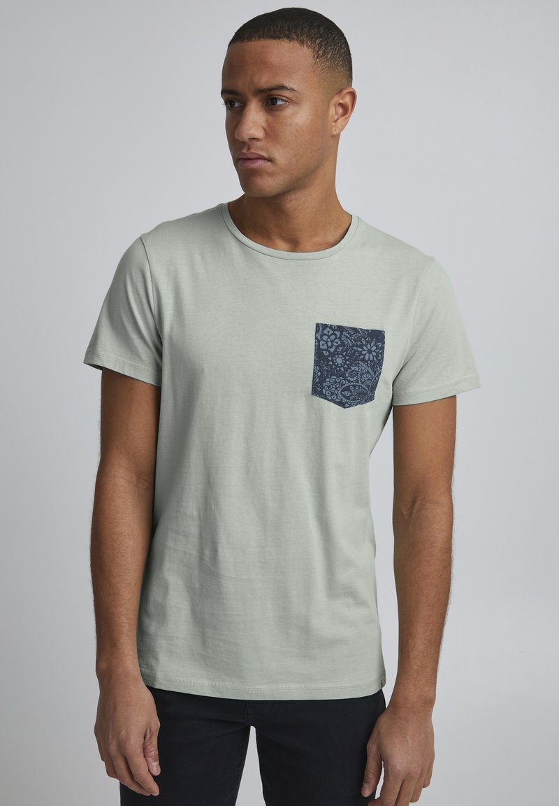 Tricou scurt din bumbac, de culoare verde deschis, cu mâneci scurte, având o buzunar cu un model bleu închis. Design clasic cu guler rotund. Textură netedă, fără aplicații.