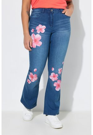 Person iført blå jeans med lyserøde blomsterprint, en orange top og hvide sneakers, stående foran en ensfarvet hvid baggrund.