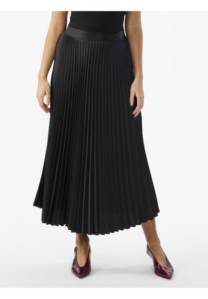 Femme portant une longue jupe noire plissée et des chaussures à talons hauts pointues rouge foncé, debout devant un fond blanc.