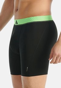 Pánský torzo v černých těsných šortkách se světle zeleným pasem, na kterém je černé logo na bílém pozadí.