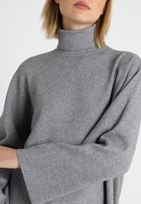 Pull à col roulé gris avec une texture douce, des manches larges et une coupe décontractée, avec un col sans couture. Le tissu semble léger.
