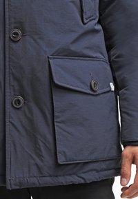 Granatowa parka o teksturowym wykończeniu, z dużą kieszenią z przodu, zapięciami na guziki i przeszyciami wzdłuż szwów.