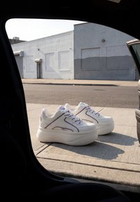 Weiße Plateau-Sneaker mit lila Schnürsenkeln und Details, die auf einem Betonweg neben einer Autotür im urbanen Straßenumfeld liegen.