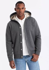 Grå hoodie med blødt interiør, fortil knapper og foret hætte. Båret over en hvid skjorte med krave, parret med lyseblå jeans.