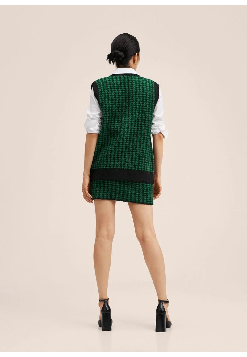 Femme debout de dos, portant un gilet et une jupe à motif pied-de-poule vert et noir, une chemise blanche et des chaussures à talons noires.