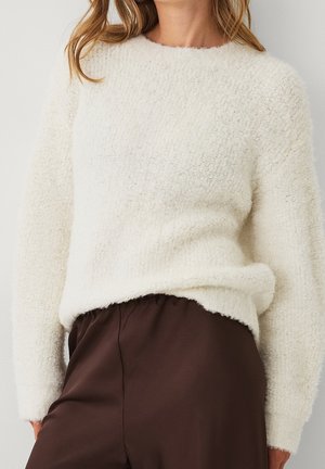 Femme portant un pull crème texturé à manches longues associé à un pantalon taille haute marron foncé sur un fond neutre.