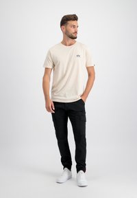 Cremfarbendes Baumwoll-T-Shirt mit kleinem Logo auf der Brust, kombiniert mit schwarzen Cargo-Hosen mit Taschen und weißen Sneakern. Einfaches Design.