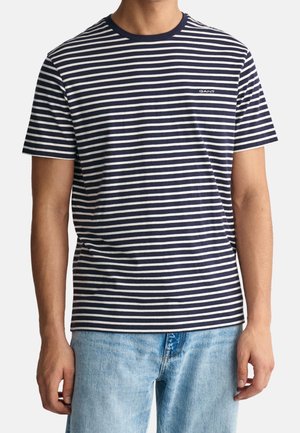 Mężczyzna ubrany w granatowo-biały t-shirt w poziome paski z krótkim rękawem i jasnoniebieskie dżinsy.