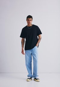 Black cotton t-shirt, relaxed fit, paired with light blue denim jeans. Notable tattoos on arms, black and white sneakers. Minimal background.