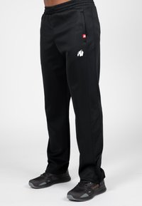 Zwarte sportbroek met een getextureerd zijpaneel, elastische tailleband en een wit gorillalog op de dij. Gekleed met zwarte sneakers.