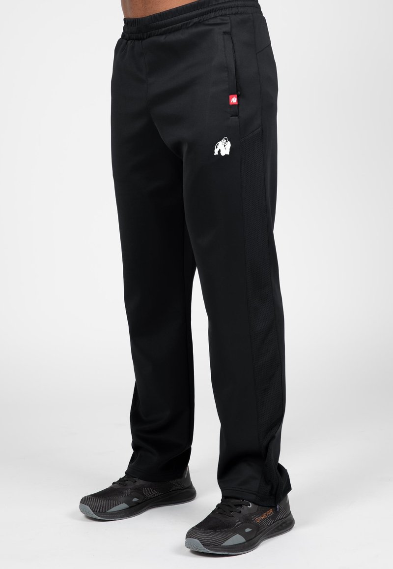 Zwarte sportbroek met een getextureerd zijpaneel, elastische tailleband en een wit gorillalog op de dij. Gekleed met zwarte sneakers.