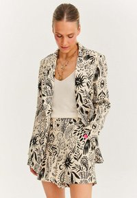 Vzorovaný dvoudílný oblek obsahující střihovou sako a šortky z béžové látky, zdobený černými ilustracemi rostlin a symbolů.