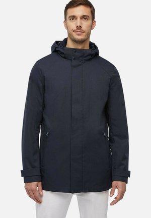 Giacca impermeabile blu navy con cappuccio, chiusura zip, tasche laterali e polsini regolabili. Texture liscia e design pulito.