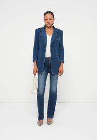 Jeans jakna v temno modri barvi z oblikovanimi rameni, gumbnim zapiranjem in dvema sprednjima žepoma, kombinirana s svetlejšim zgornjim delom, oprijetimi kavbojkami in petkami.
