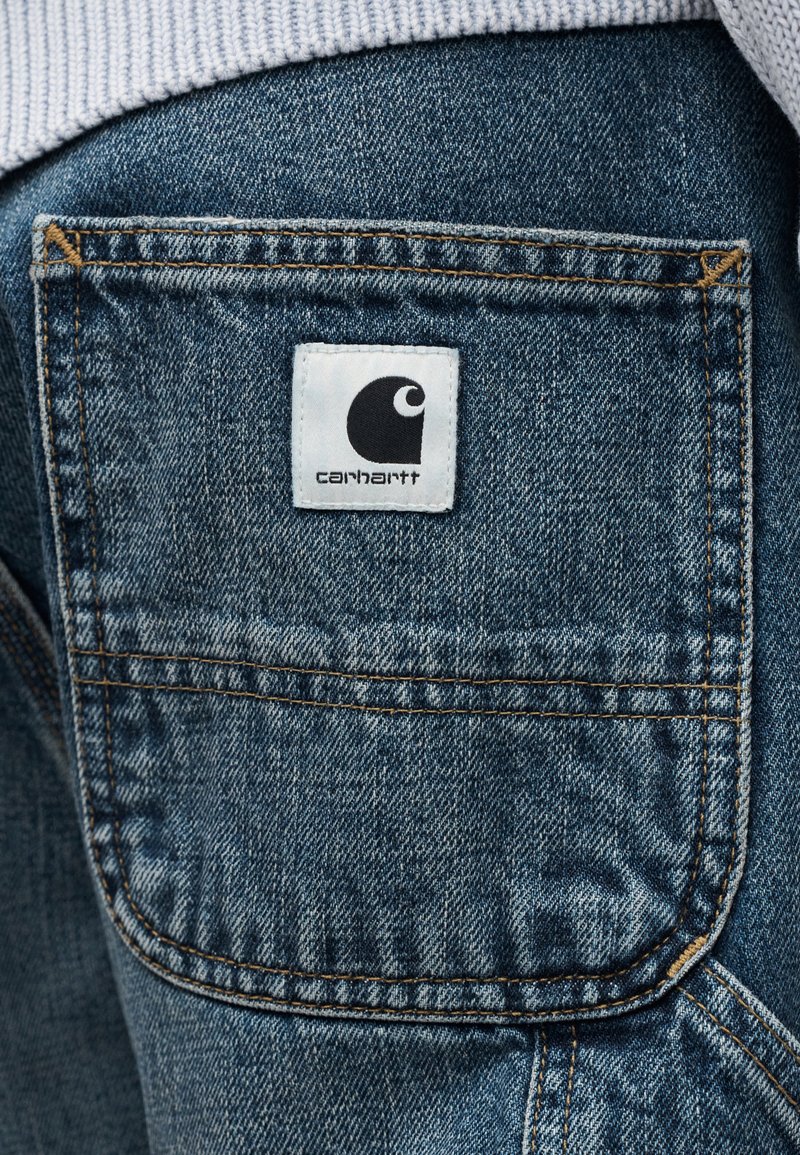 Jean en denim bleu avec une poche arrière plaquée ornée d'une étiquette blanche portant le logo Carhartt noir et des coutures contrastantes orange.