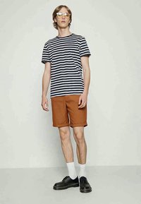 Jovem vestindo uma t-shirt listrada em preto e branco, calções de cor rust, meias brancas, sapatos pretos e óculos com lentes amarelas, de pé contra uma parede simples.