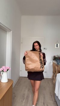 Brūna papīra soma ar melnu "zalando" logotipu, ko tur persona tumšā džemperī un svārkos, stāvot minimālistiskā iekštelpu telpā.